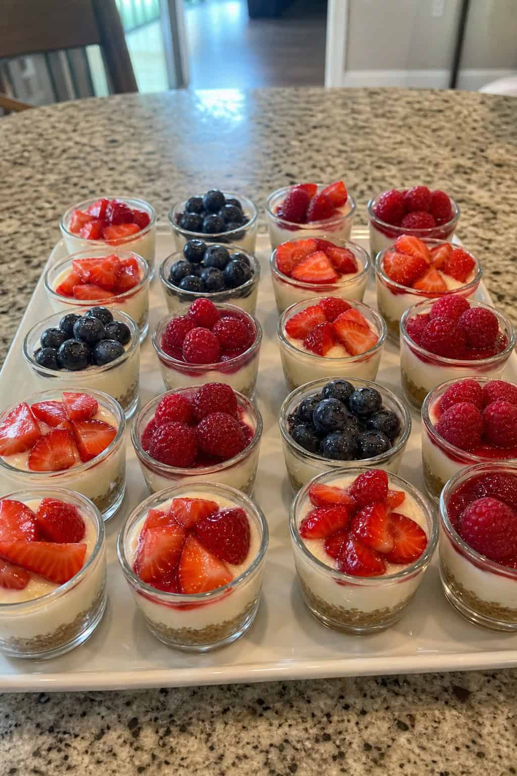 Twenty-two mini cheesecake trifles in clear cups arranged on a wooden table, topped with fresh berries and mint leaves.