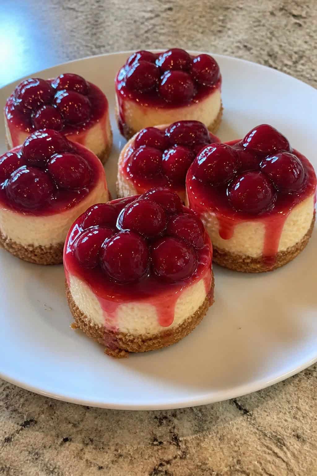 Close-up of several mini cherry pie cheesecakes topped with cherries and sauce on a wooden serving board with scattered cherries and mint leaves.