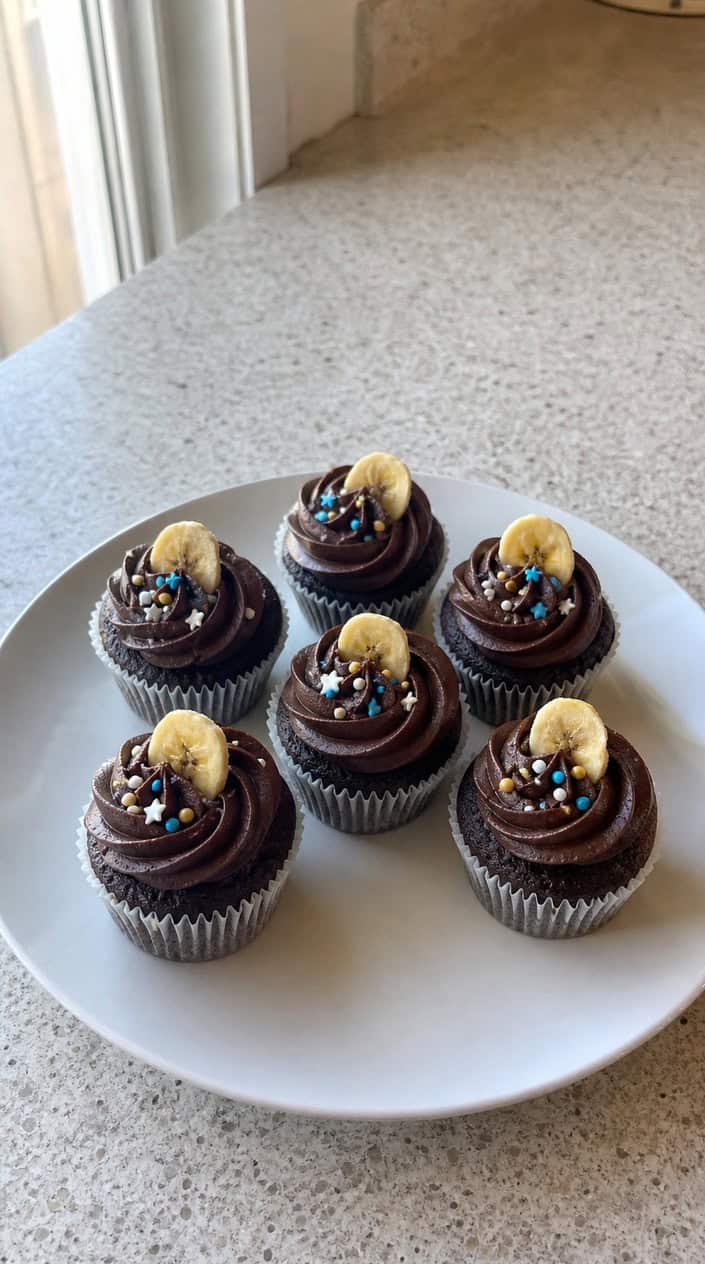 Close-up of mini chocolate banana cupcakes with chocolate frosting and banana slices on a wooden table, with soft natural lighting and a blurred background.
