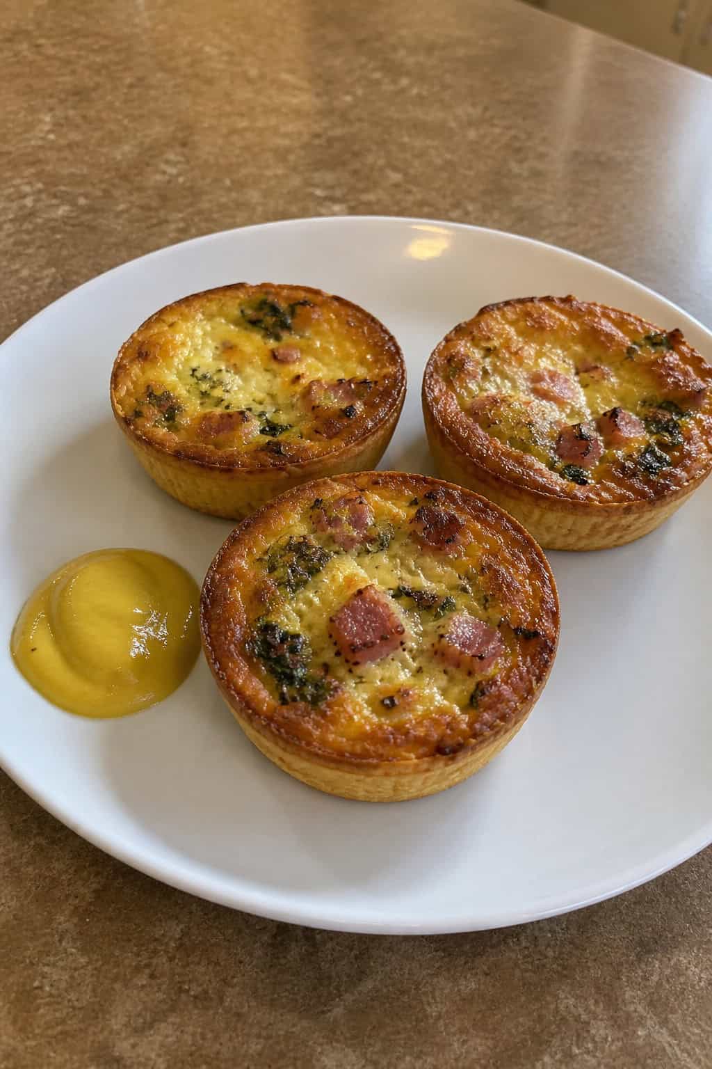 Three mini quiches on a white plate served with a small bowl of mustard on a table.
