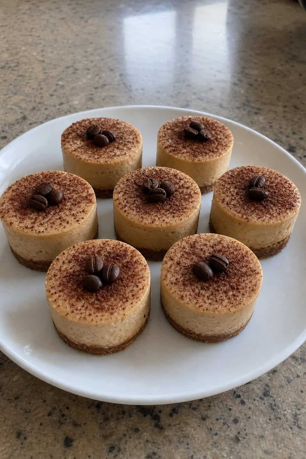 A plate of mini espresso cheesecakes garnished with coffee beans and cocoa powder on a wooden table with a cozy party setting in the background.