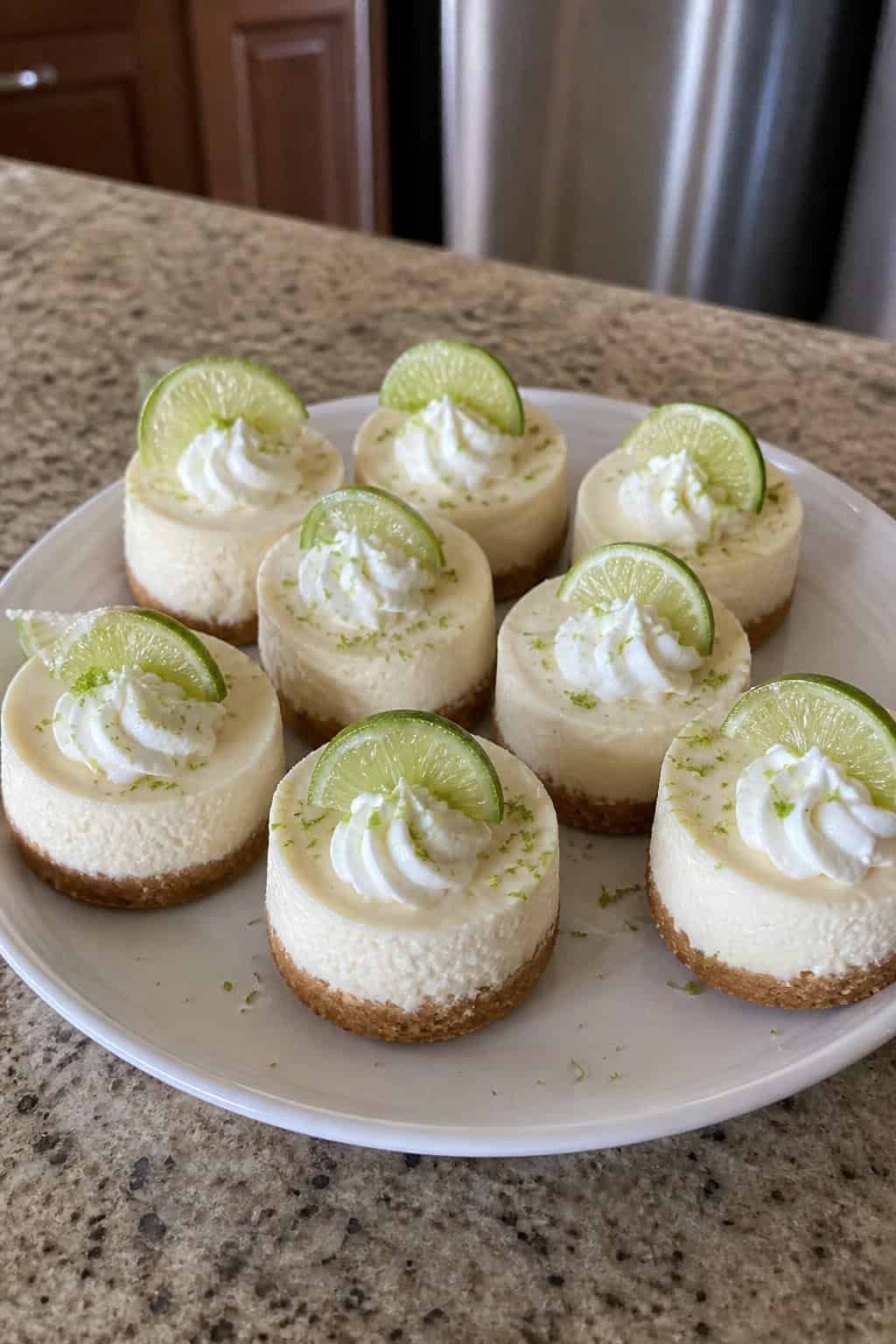A wooden board with several mini key lime cheesecakes topped with lime slices and whipped cream, surrounded by fresh limes and mint leaves.