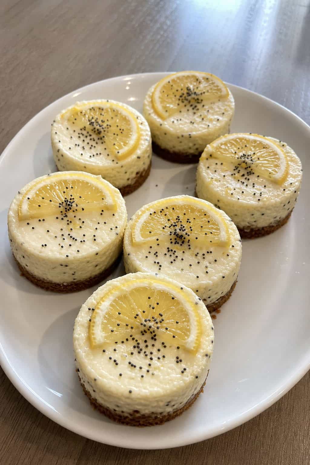 Several mini lemon poppy seed cheesecakes arranged on a white platter with lemon slices and poppy seeds, placed on a wooden table with fresh lemons and mint leaves nearby.