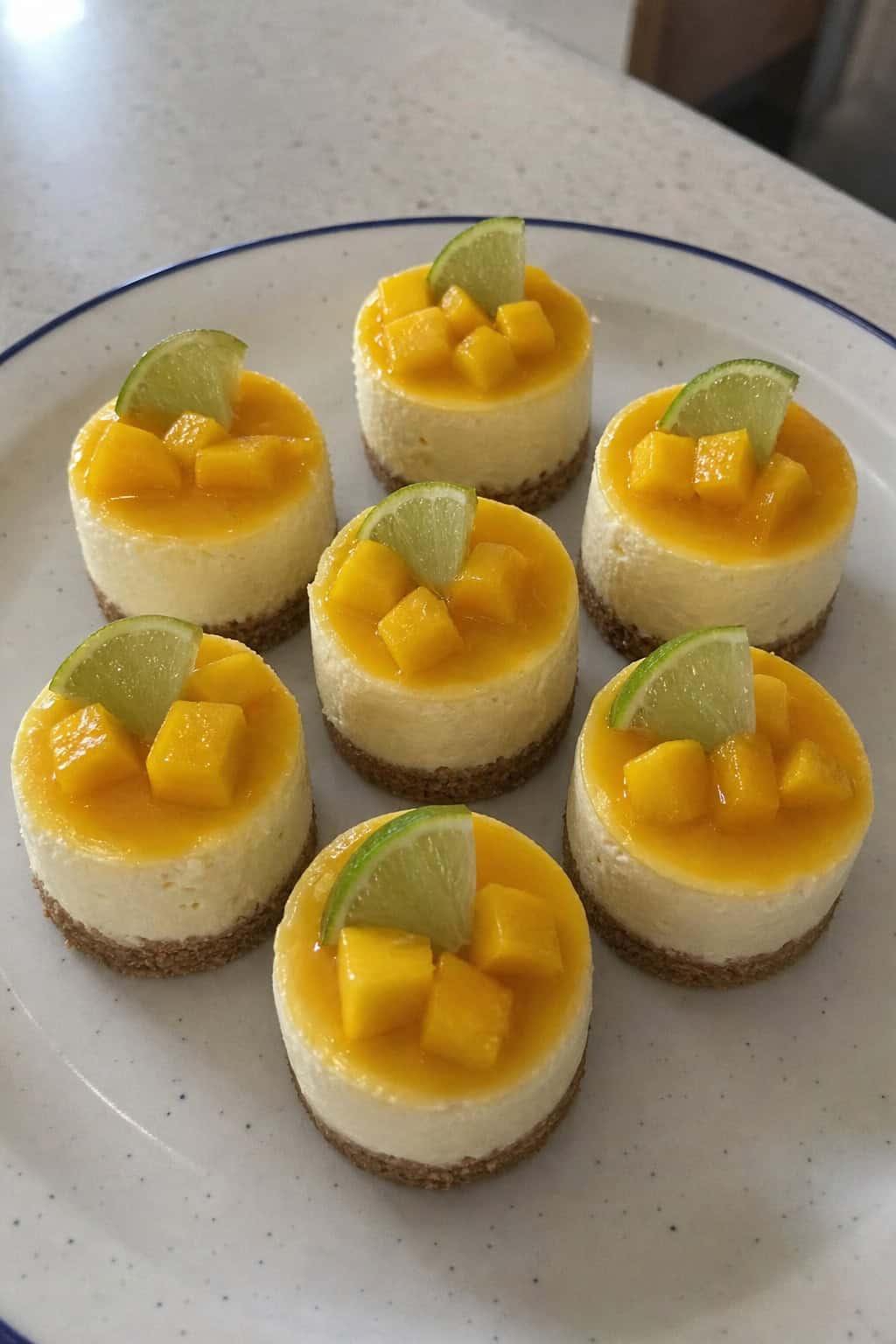 Close-up of several mini mango lime cheesecakes on a wooden board, garnished with lime slices and fresh mango, with some mint leaves around.