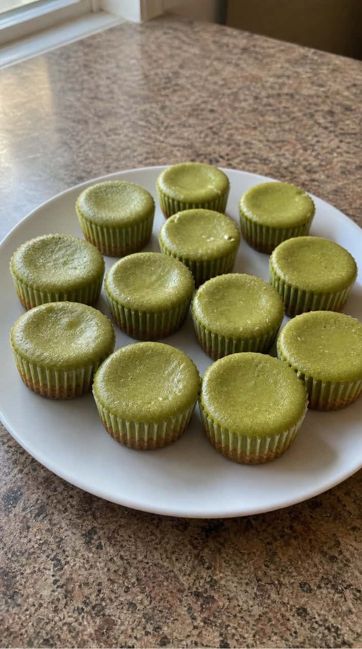 Six small round matcha cheesecakes on a dark wood serving board, their pale green surfaces dusted with powdered sugar and each garnished with a single small mint leaf.