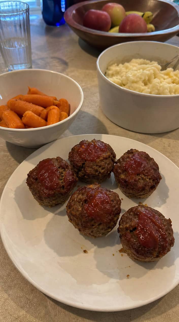 Mini meatloaf muffins on a wooden board with fresh herbs and colorful kid-friendly side dishes on a table.