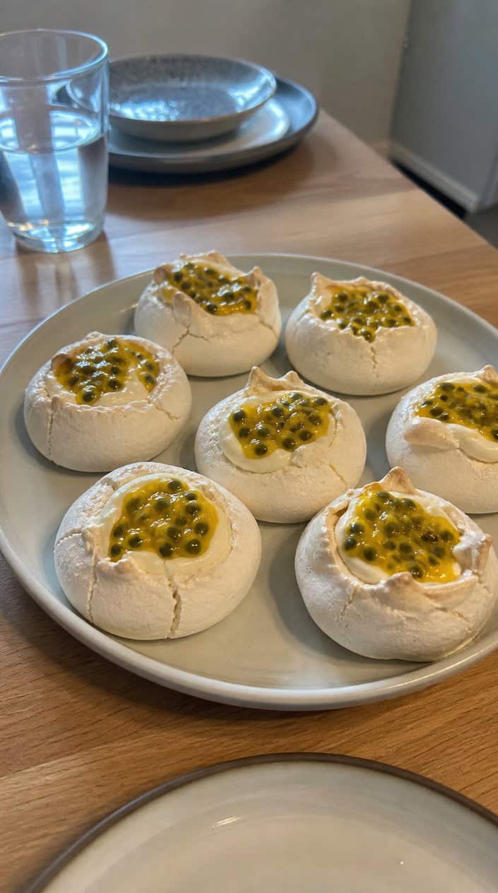 A close-up of six small meringue nests on a parchment-lined surface, each filled with passionfruit pulp and seeds, surrounded by halved fresh passionfruits and a few mint leaves.