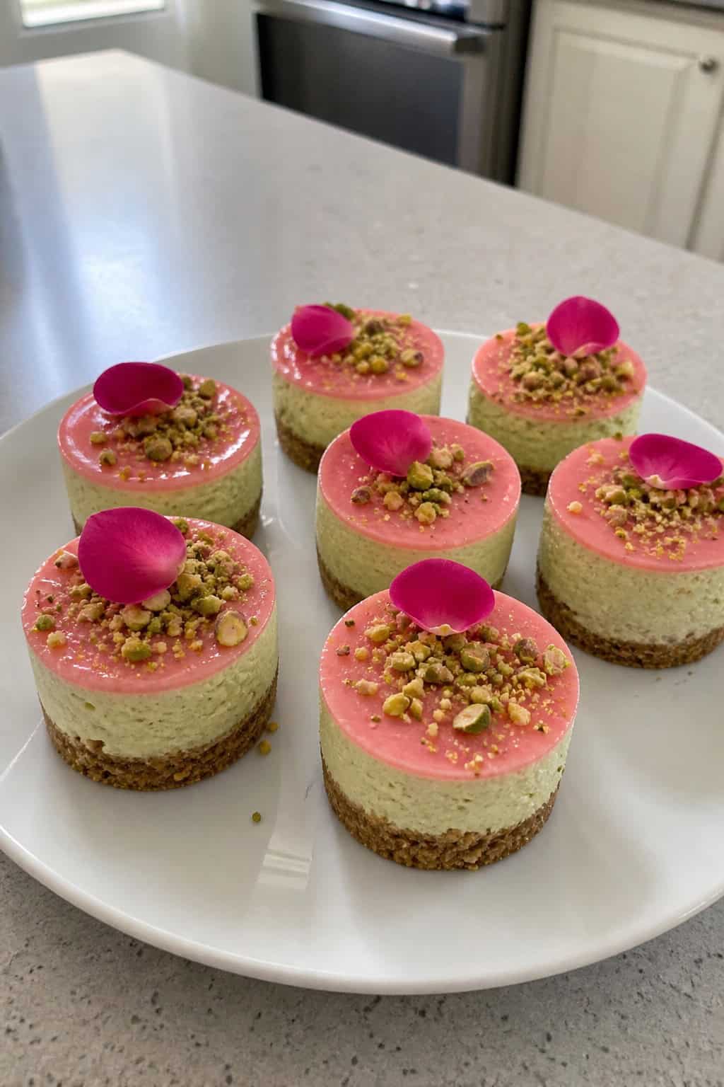 A platter of small pistachio and rosewater cheesecakes garnished with crushed pistachios and rose petals on a wooden table with party decorations in the background.