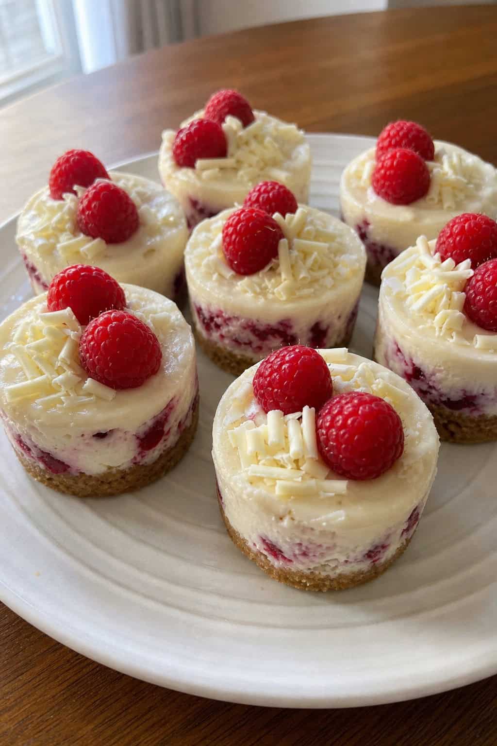 Close-up of mini white chocolate raspberry cheesecakes topped with fresh raspberries and white chocolate shavings on a wooden serving board.