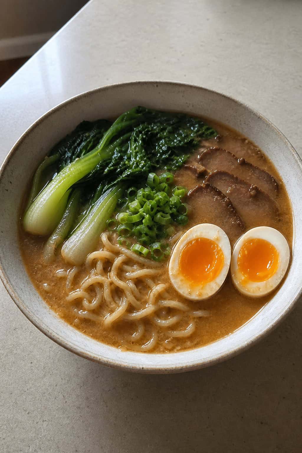 A bowl of miso ramen with soft-boiled eggs and bok choy on a wooden table, with chopsticks and a small dish nearby.