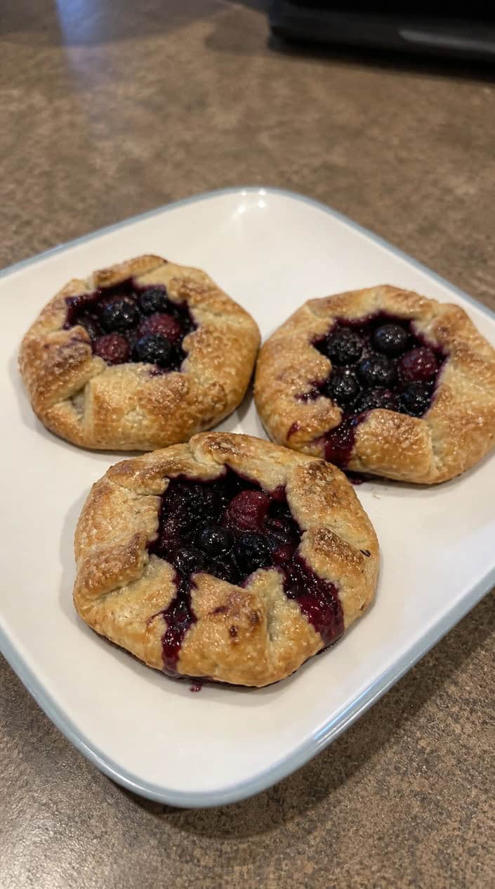 Freshly baked mixed berry galette hand pies on a wooden board with fresh berries and powdered sugar.