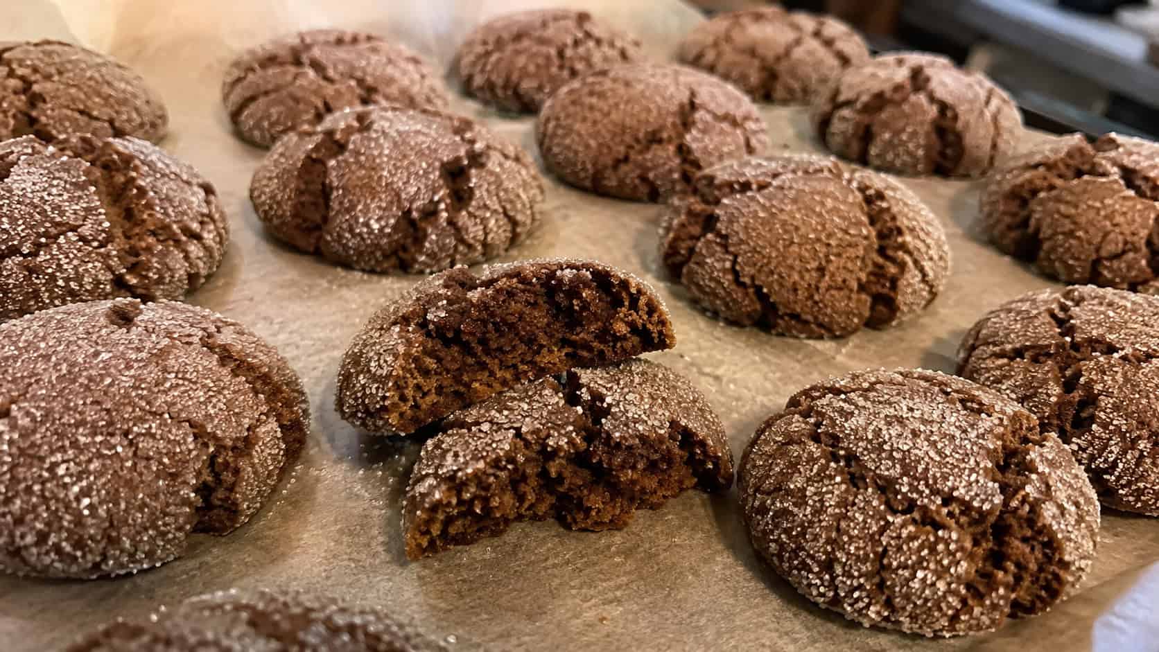 finished molasses ginger crinkle cookies on parchment, dark brown cookies with cracked crinkled sugar-coated tops, one broken open showing a soft chewy center, warm natural kitchen light, casual home baking