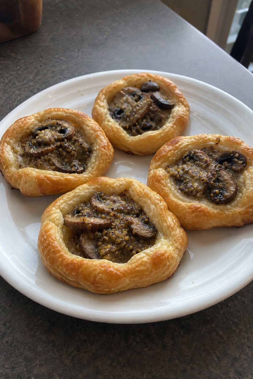 Several small mushroom and Dijon mustard puff pastry appetizers arranged on a wooden board with fresh herbs nearby.