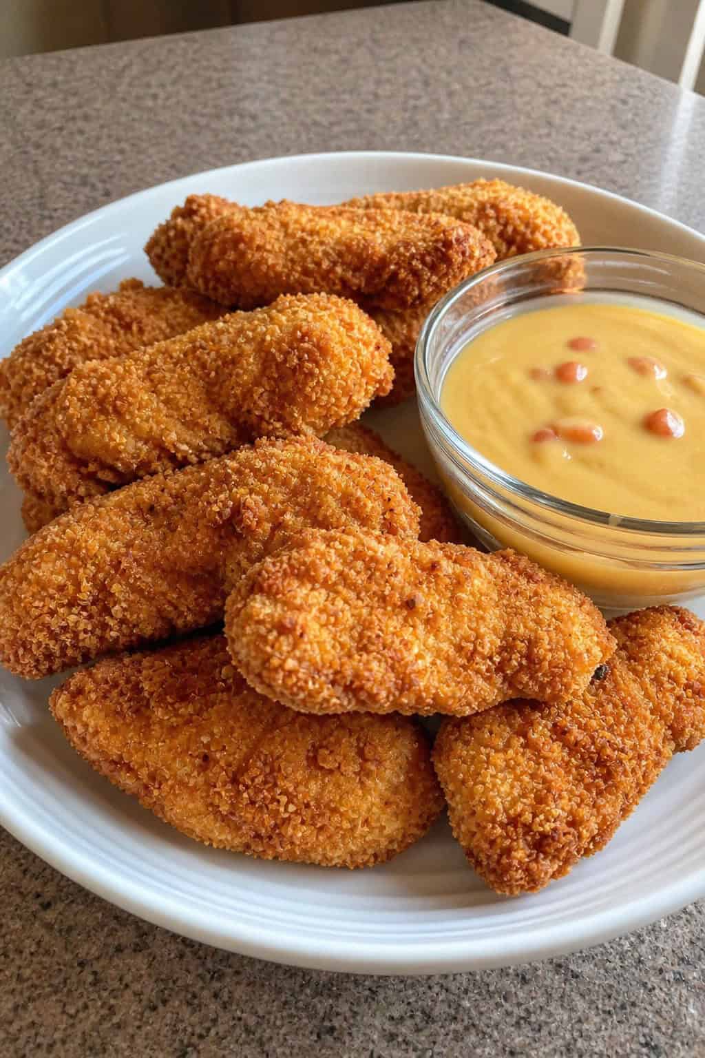 A plate of crispy nacho-style chicken tenders with a bowl of melted queso cheese on a wooden table.