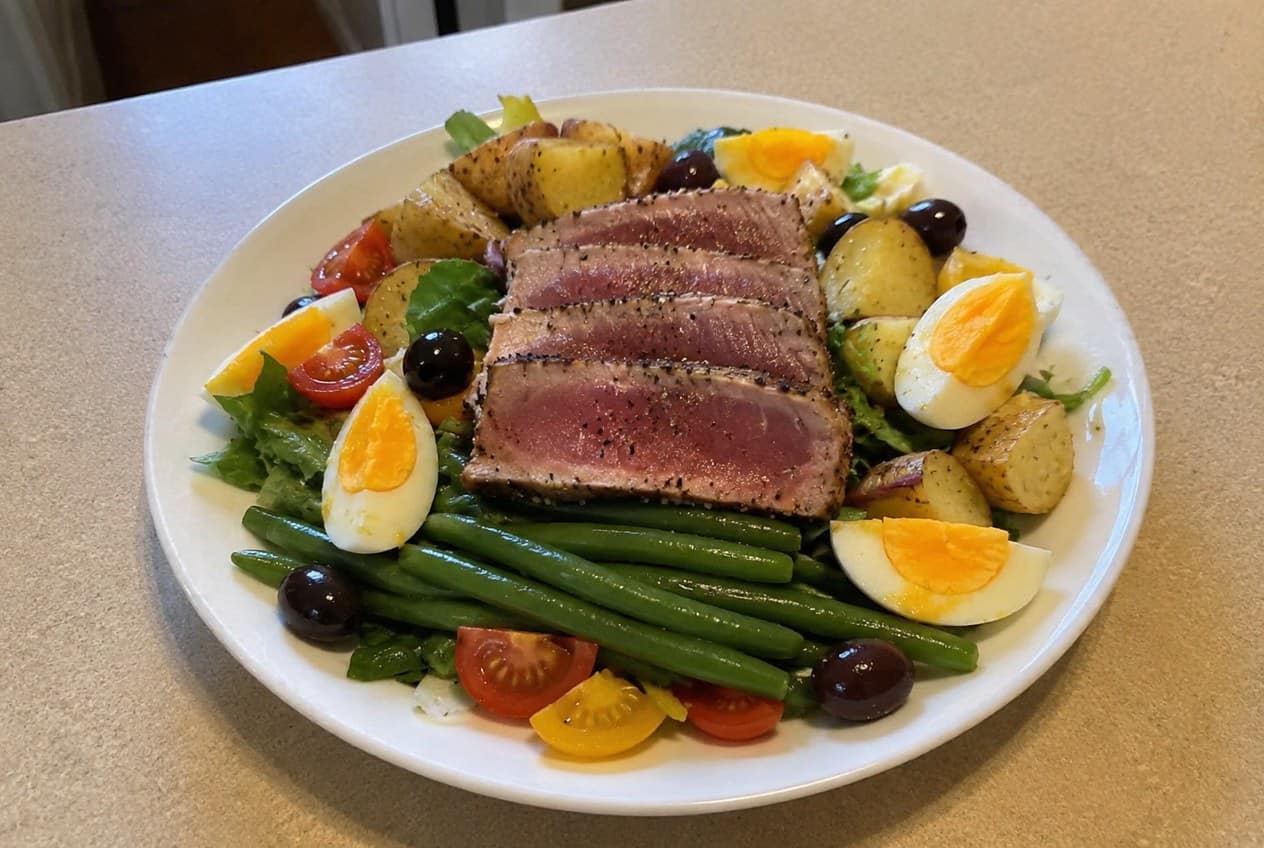 A wide plate arranged with sliced seared tuna, halved hard-boiled eggs, crisp green beans, baby potatoes, cherry tomatoes, black olives, and chopped romaine, drizzled with Dijon vinaigrette.