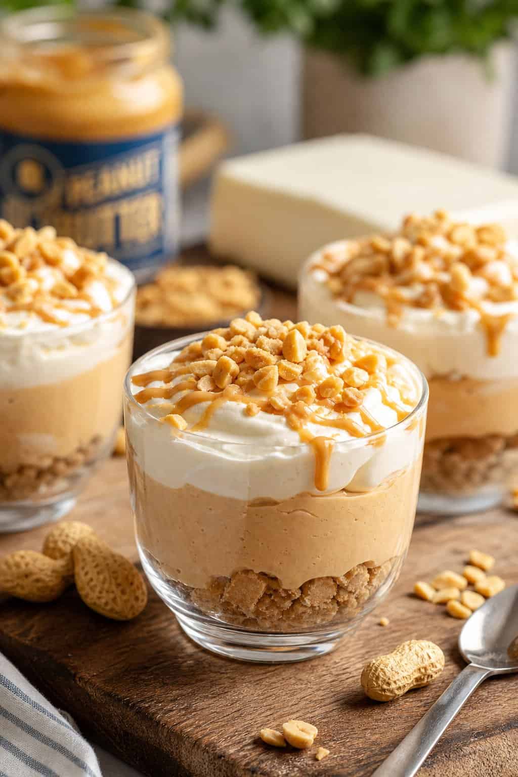 Three no-bake peanut butter cheesecake cups in short clear glasses, topped with a swirl of peanut butter drizzle and crushed salted peanuts, arranged on a wooden surface with a jar of peanut butter visible in the background.