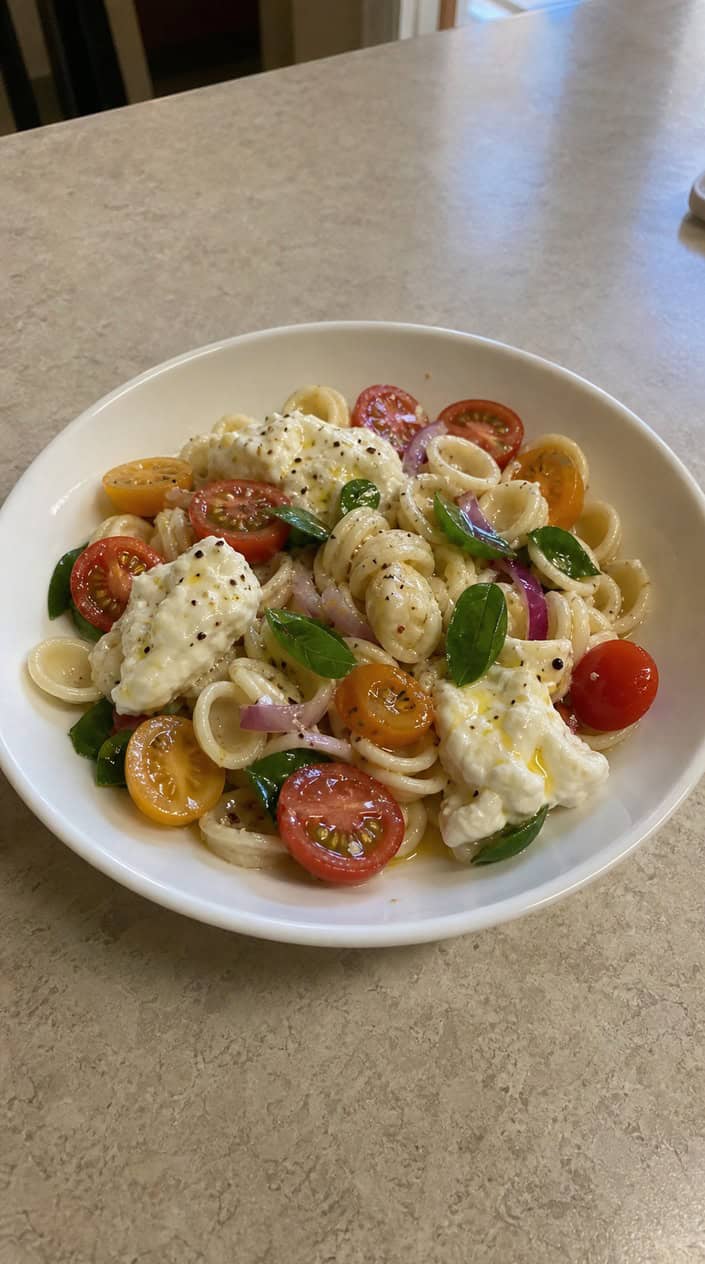 A bowl of Caprese pasta salad with cherry tomatoes, basil, and burrata cheese on a wooden table.