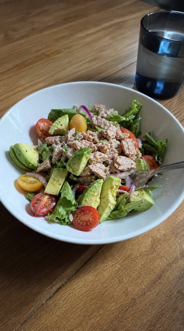 A rustic ceramic bowl filled with pale green avocado tuna salad, garnished with cilantro and served with two lime wedges on the side, set on a light wood surface.