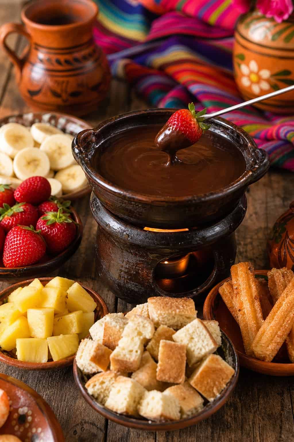 A table with a clay pot of melted chocolate fondue surrounded by fruits and pieces of bread for dipping.