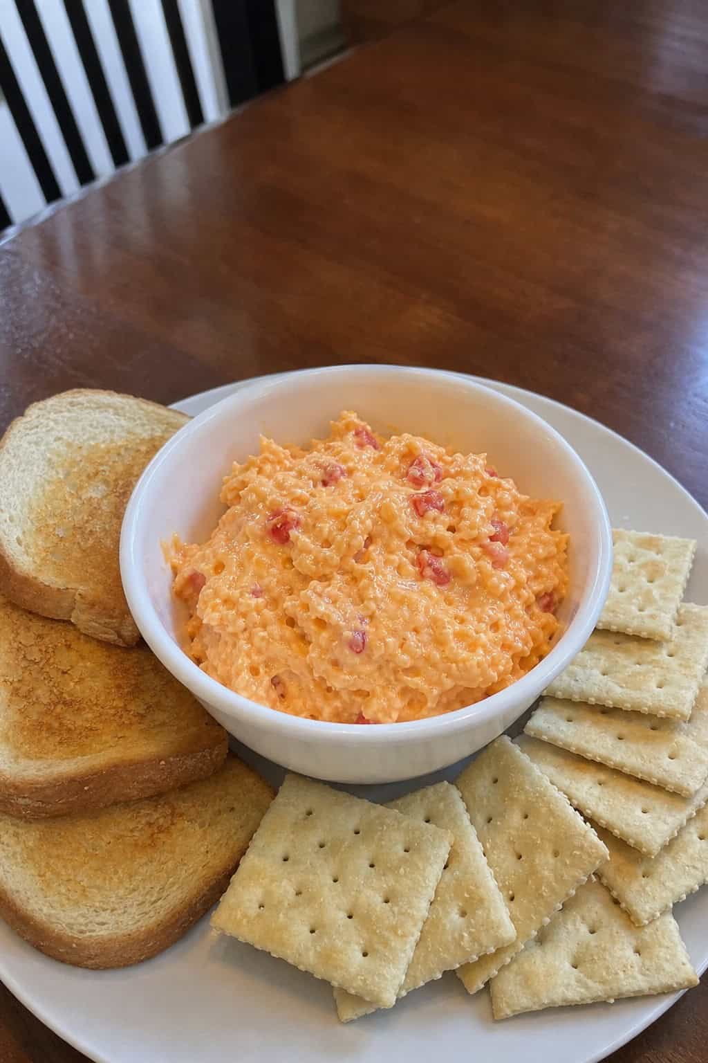 Bowl of creamy pimiento cheese spread with sharp cheddar and mayonnaise on a wooden board, surrounded by fresh ingredients and toasted bread slices.