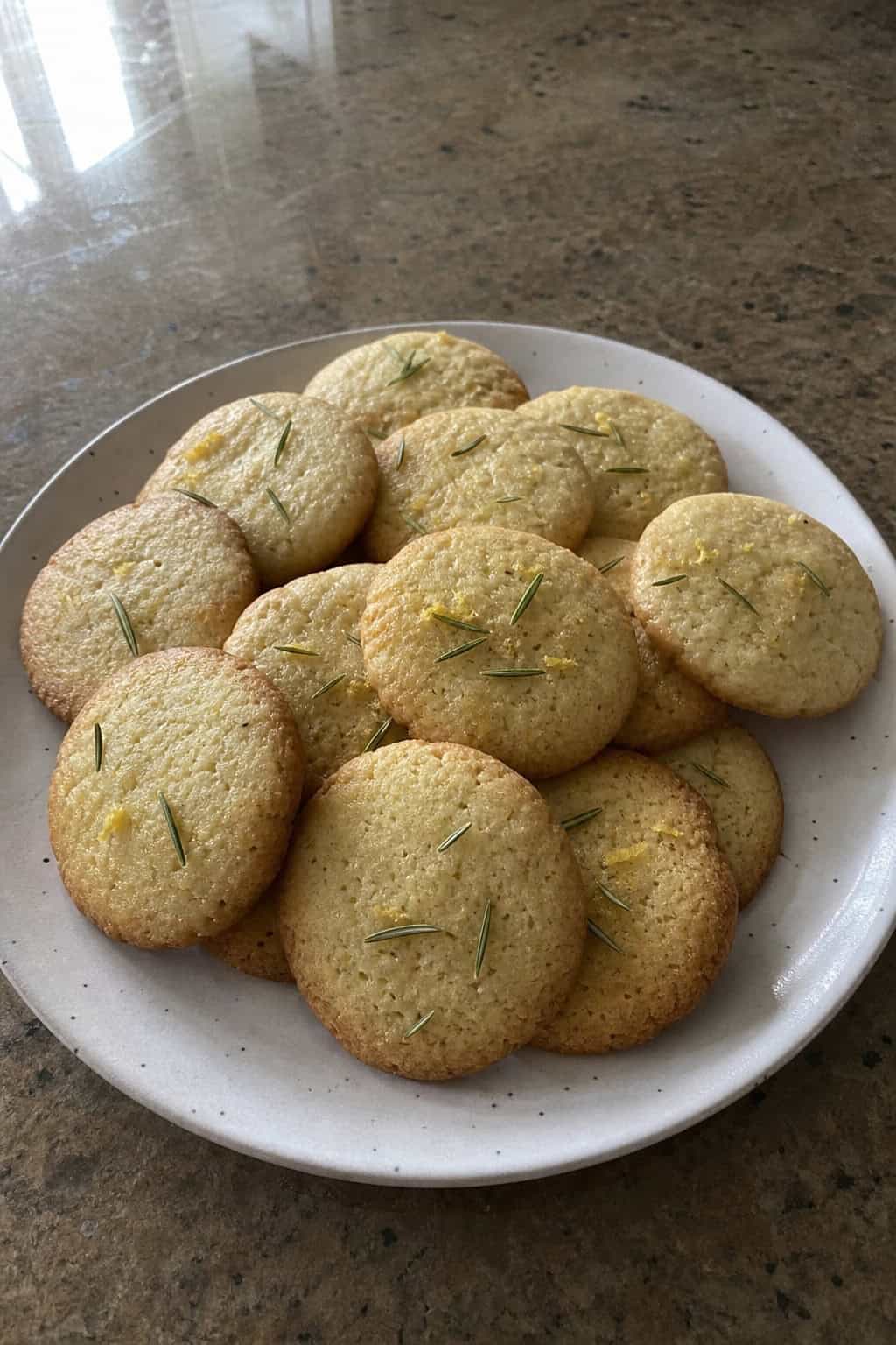 Plate of golden brown olive-oil rosemary lemon cookies on a wooden table with fresh lemons, rosemary sprigs, and a small bottle of olive oil.