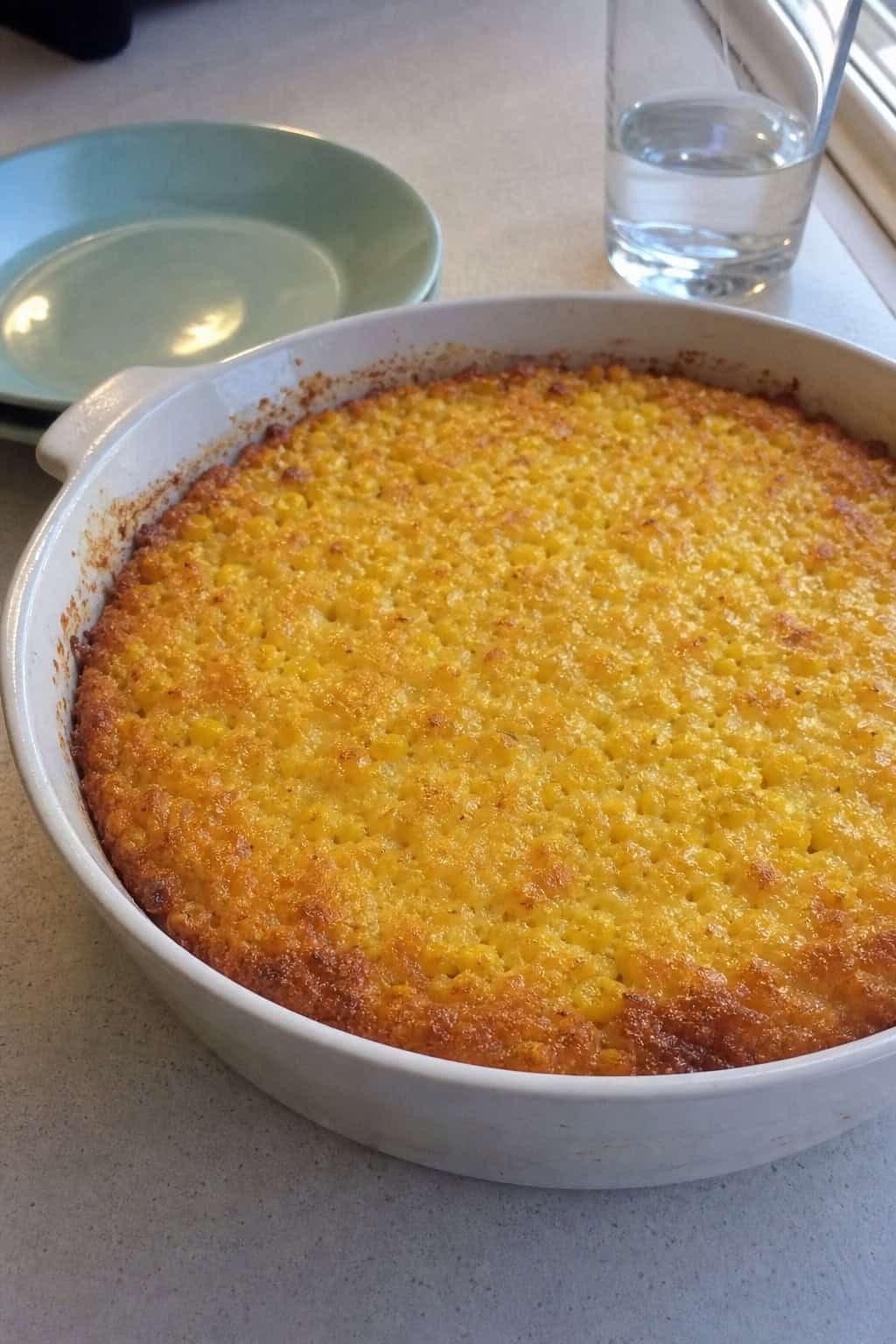 A freshly baked corn casserole in a white ceramic dish on a wooden table with a single large mixing bowl and a folded striped kitchen towel beside it.