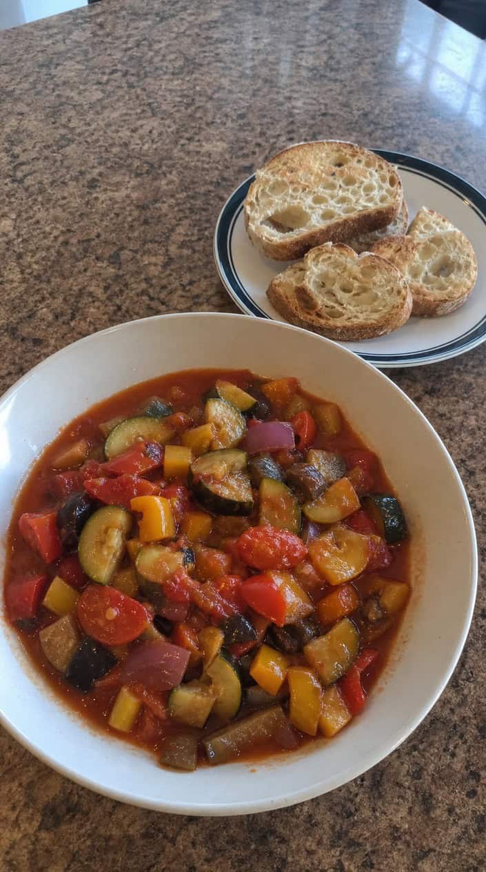 A bowl of colorful summer ratatouille with slices of crusty bread on a wooden table.