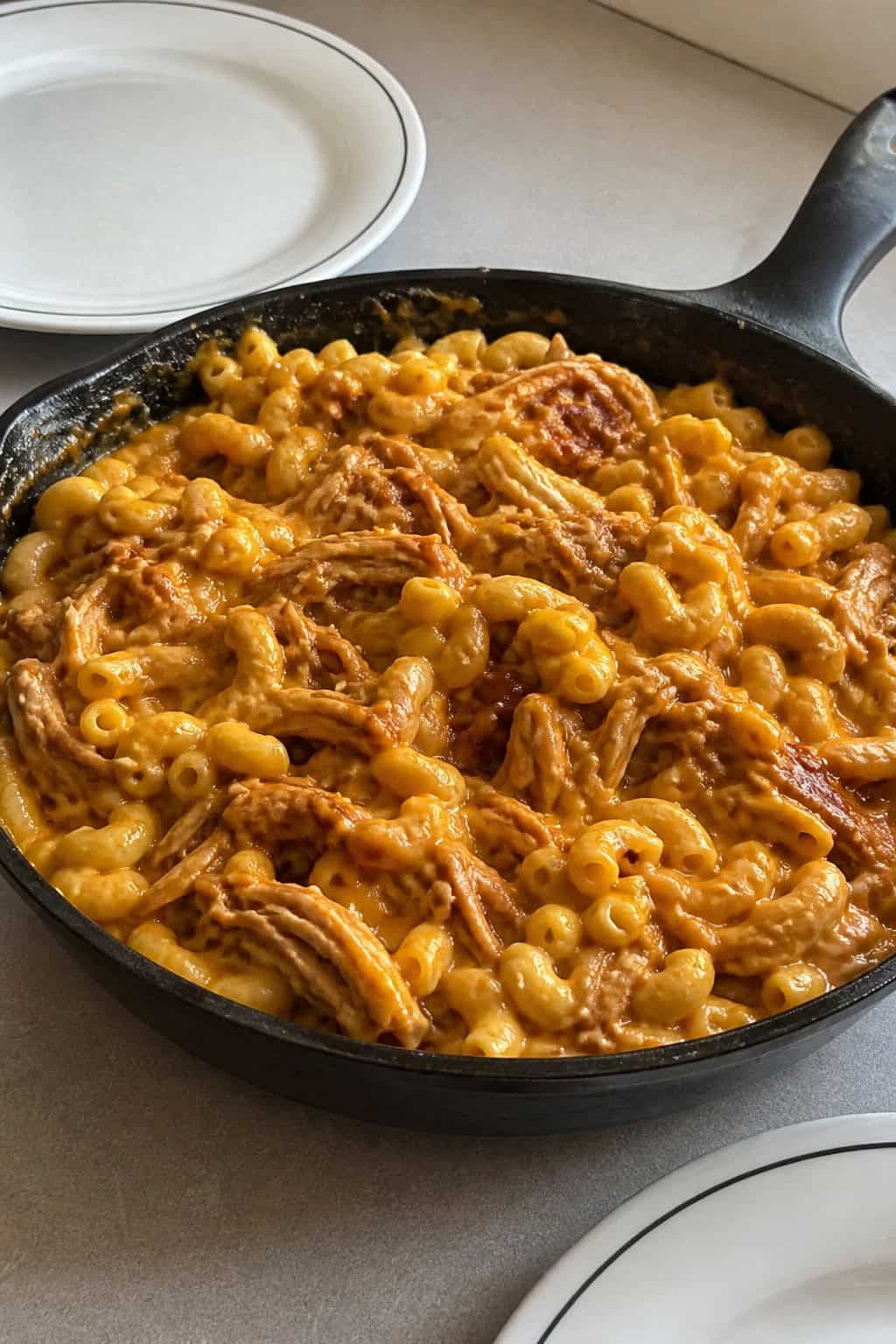A cast iron skillet of creamy elbow macaroni mixed with shredded BBQ chicken, the sauce glossy and golden-brown around the edges, garnished with fresh parsley on a wooden table.