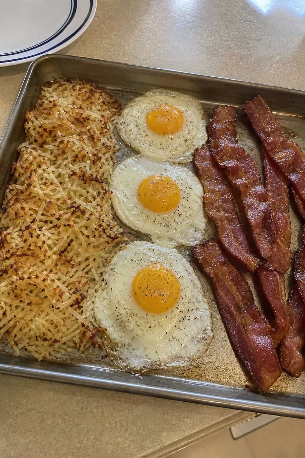 A rimmed baking sheet with a golden-brown bed of shredded hash browns, four sunny-side-up eggs with set whites and bright yolks, and crispy flat strips of bacon on a wooden table.