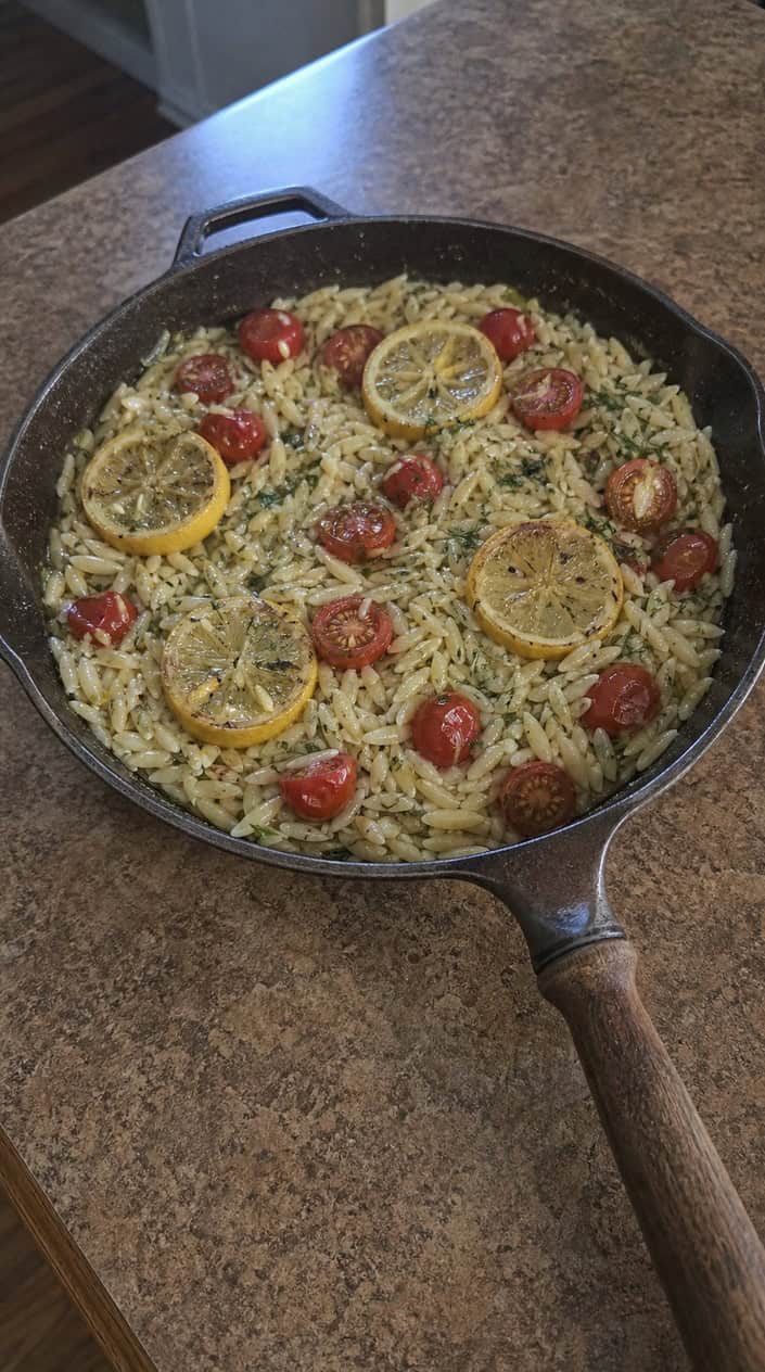 A skillet on a stovetop filled with lemon-dill orzo pasta and cherry tomatoes, with fresh ingredients around it on a kitchen counter.