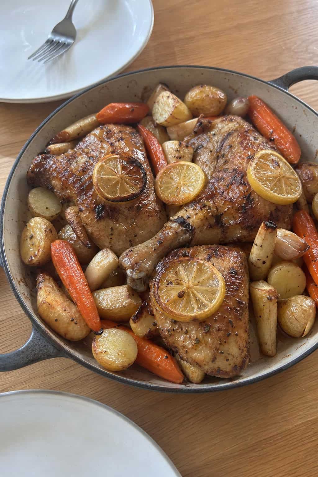 A pan with lemon garlic chicken and roasted root vegetables on a wooden table, with a jar of homemade chicken stock in the background.