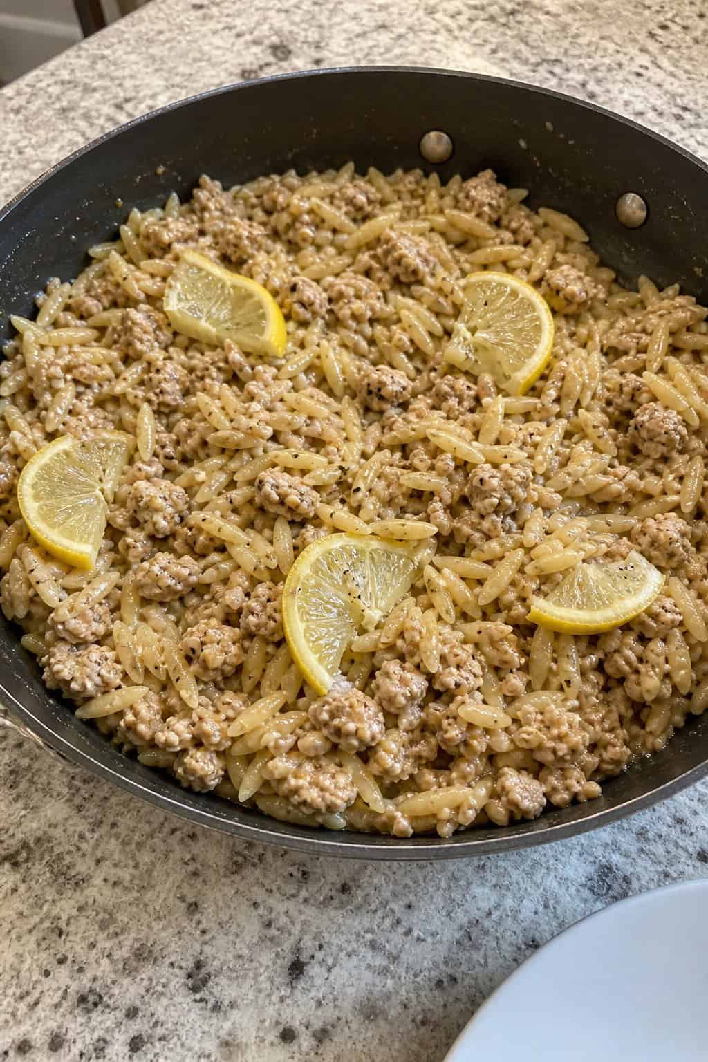 25 Quick and Easy Minced Chicken Recipes 3 A skillet filled with lemon garlic minced chicken and orzo pasta garnished with parsley and lemon slices on a wooden table.
