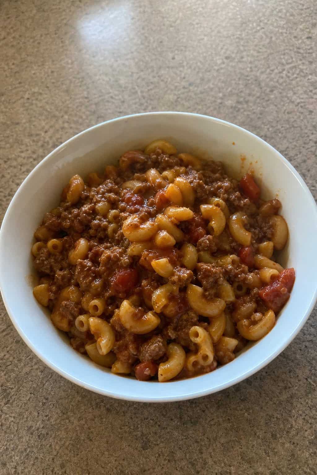 A bowl of American goulash with ground beef, elbow macaroni, and tomato sauce garnished with parsley on a wooden table.