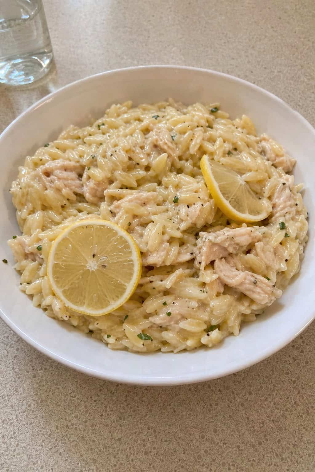 A bowl of lemon garlic canned chicken orzo garnished with lemon slices and parsley on a wooden table with fresh ingredients around it.