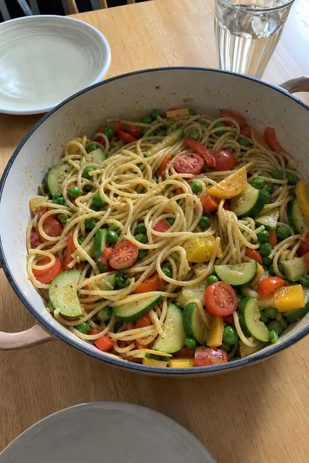 A pot of colorful pasta primavera with fresh vegetables on a wooden table, surrounded by vegetable peels and kitchen tools.