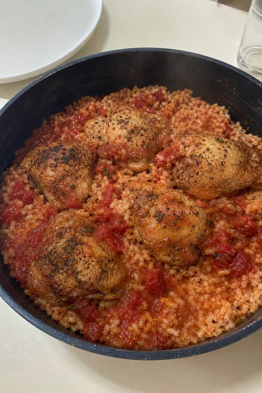 A wide skillet on a wooden table filled with chicken thighs half-submerged in a deep red tomato basil sauce over rice, topped with scattered fresh basil leaves and a few whole tomatoes visible at the edge.