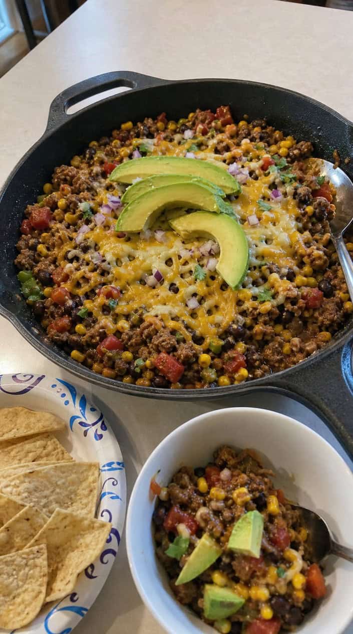 A skillet filled with a colorful turkey taco mixture including beans, corn, tomatoes, and avocado on a wooden table.