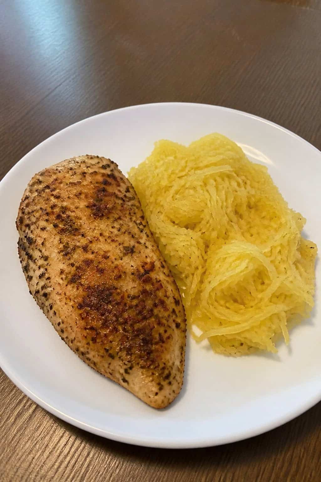 A plate with ranch seasoned chicken and roasted spaghetti squash on a wooden table with fresh herbs and a bowl of dressing in the background.