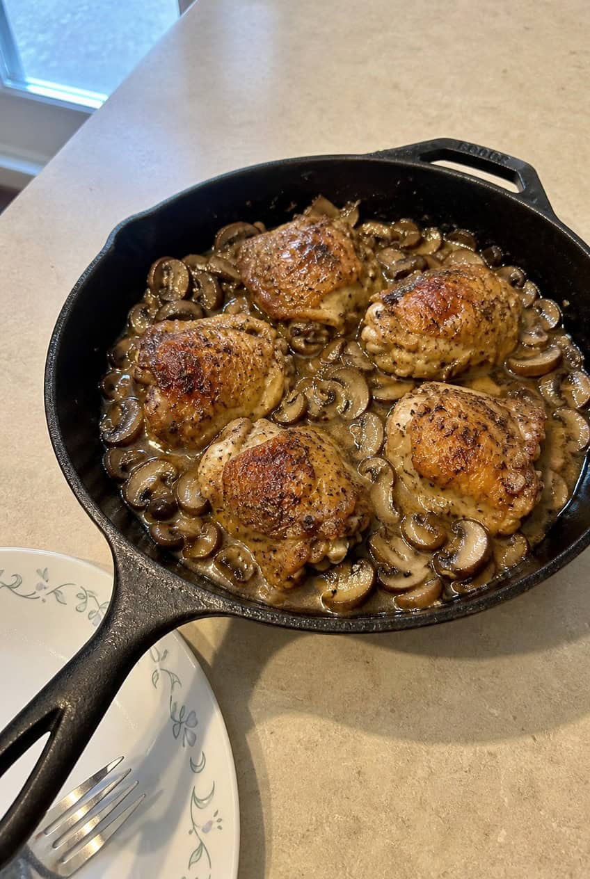 A large skillet with seared chicken breasts and sliced brown mushrooms in a creamy pale pan sauce, garnished with fresh thyme leaves on a wooden surface.