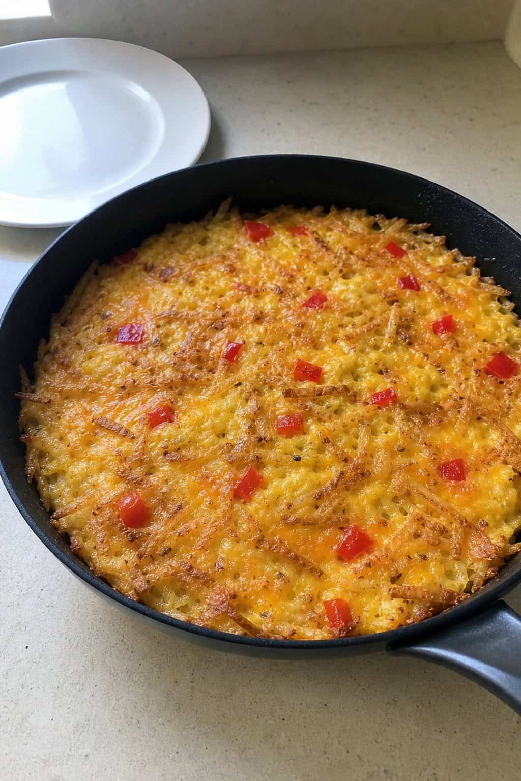 A cast-iron skillet filled with a golden hashbrown casserole topped with melted cheese, diced vegetables, and egg, sitting on a kitchen countertop with a glass of orange juice and a bowl of fresh fruit nearby.