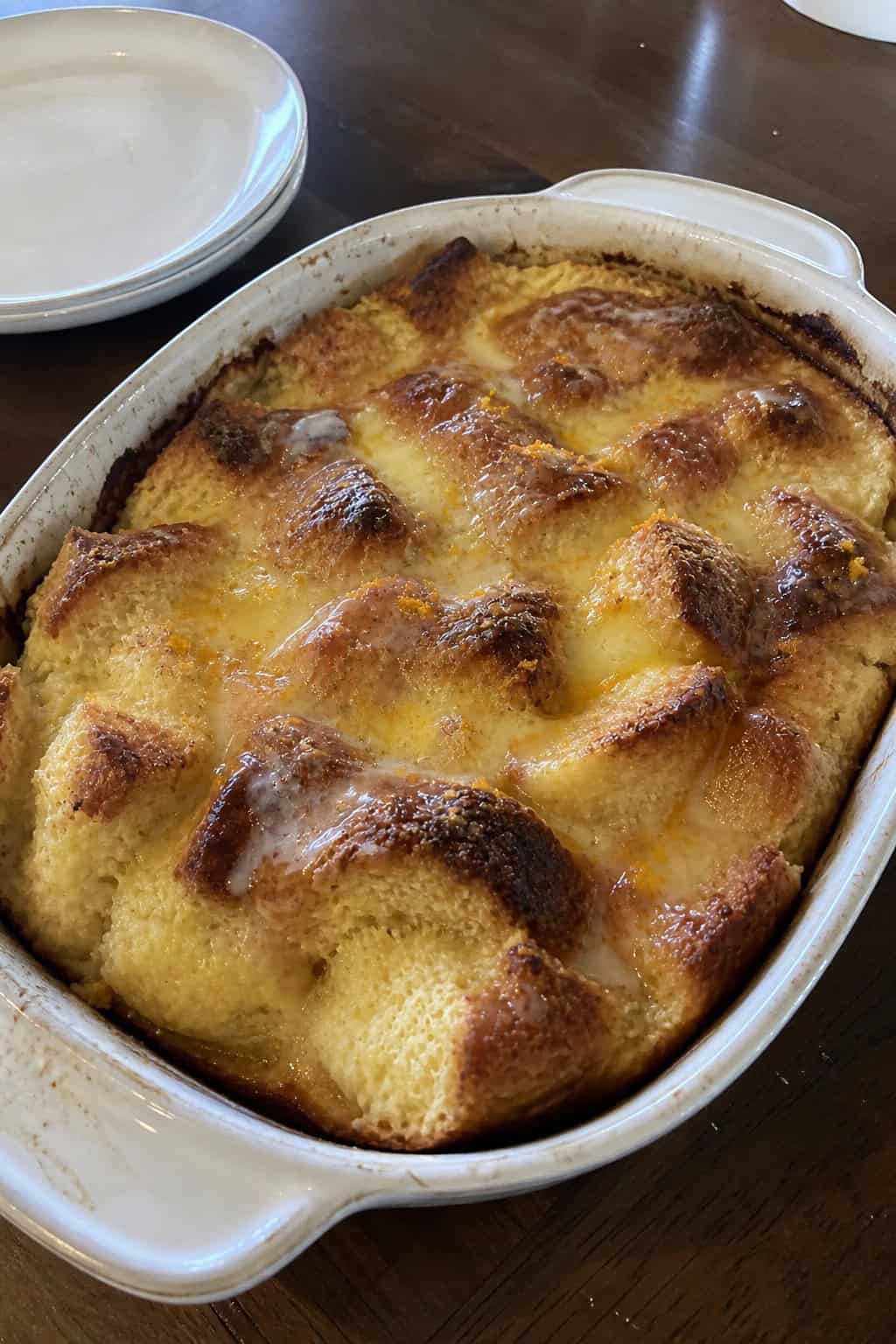 A baked orange-glazed ricotta French toast casserole in a white ceramic dish with a glossy powdered sugar glaze drizzled across the top, with fresh orange slices and zest arranged alongside on a wooden table.