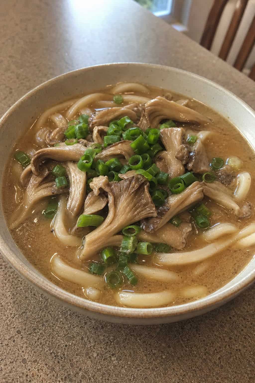 A steaming bowl of oyster mushroom and miso broth with udon noodles on a wooden table, garnished with scallions and mushrooms.