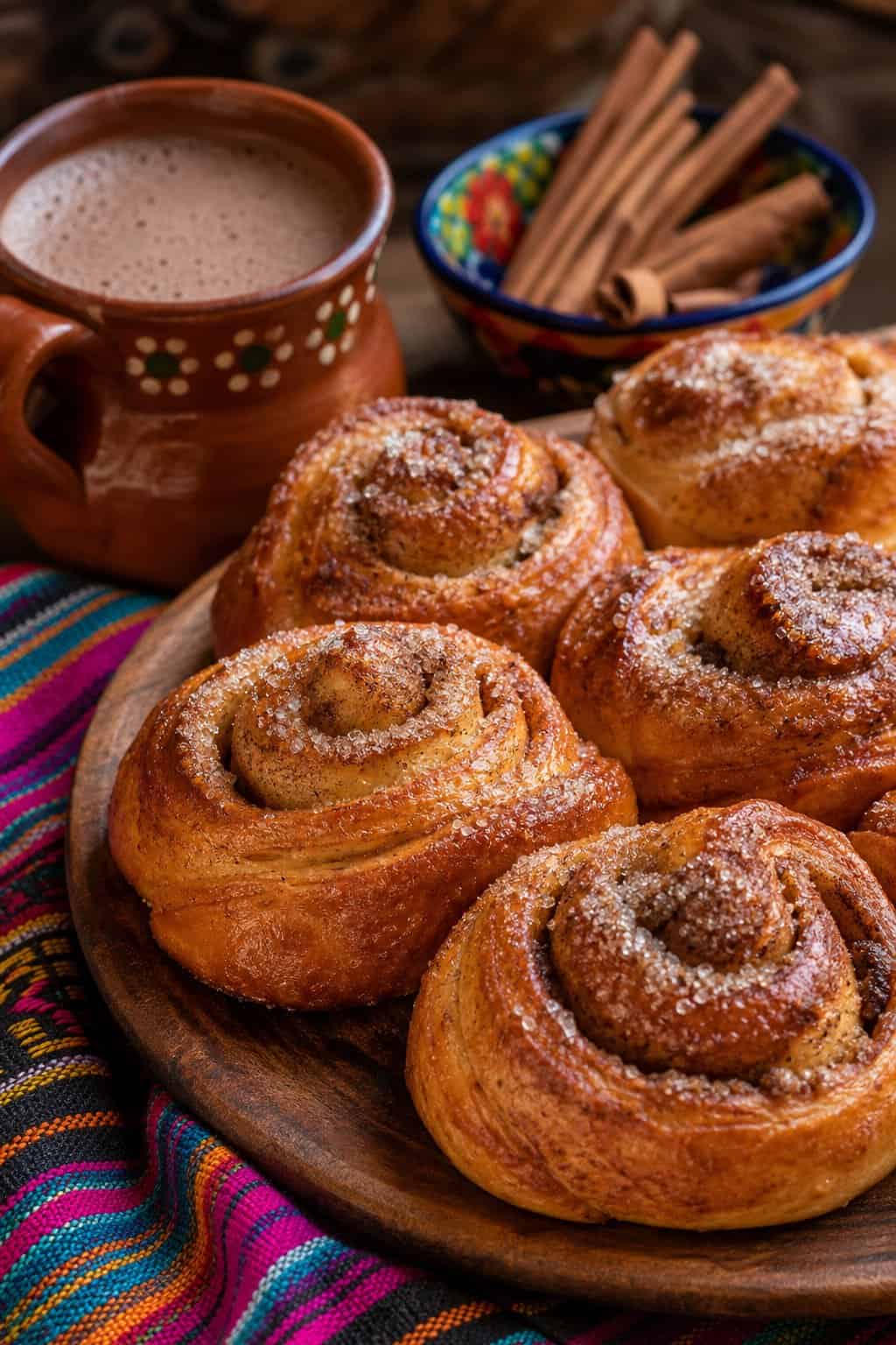 Close-up of freshly baked pan dulce cinnamon rolls on a wooden platter with cinnamon sticks and a cup of hot chocolate.
