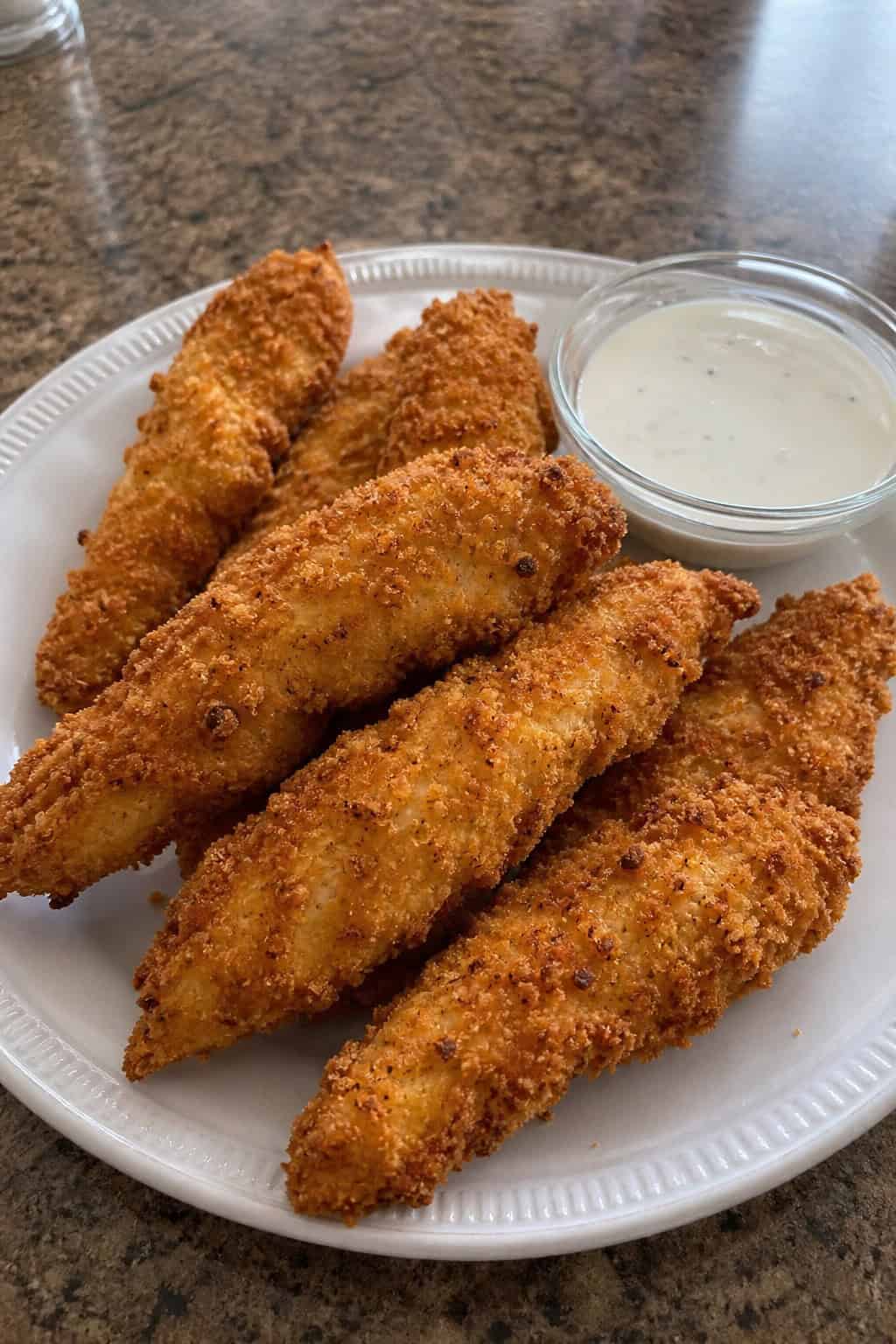 Crispy panko-crusted chicken tenders on a white plate with a bowl of ranch dipping sauce and lemon wedges on a wooden table.