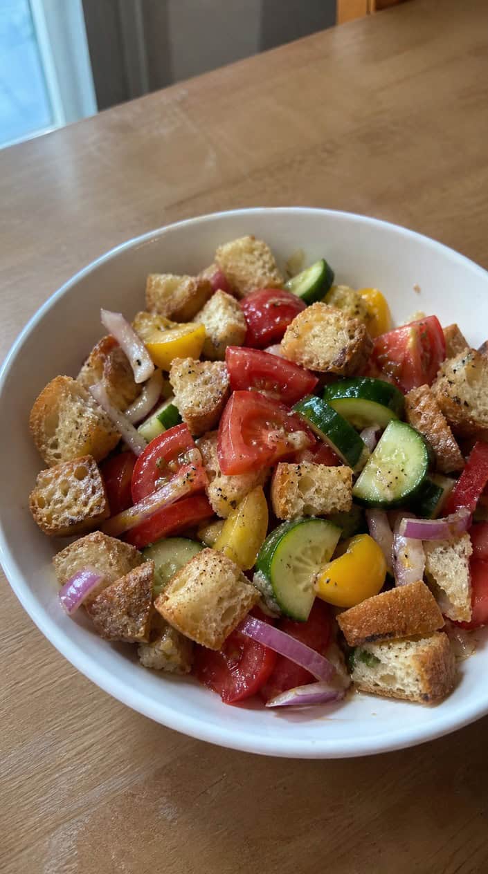 A bowl of panzanella salad with bread cubes, ripe tomatoes, basil, and onions on a wooden table with fresh ingredients around it.