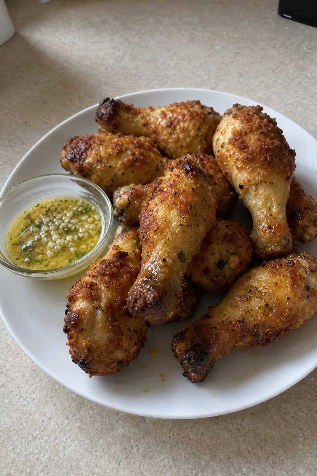 Golden parmesan-crusted chicken drumsticks on a white plate garnished with chopped flat-leaf parsley, with a small ramekin of garlic butter sauce on the side.