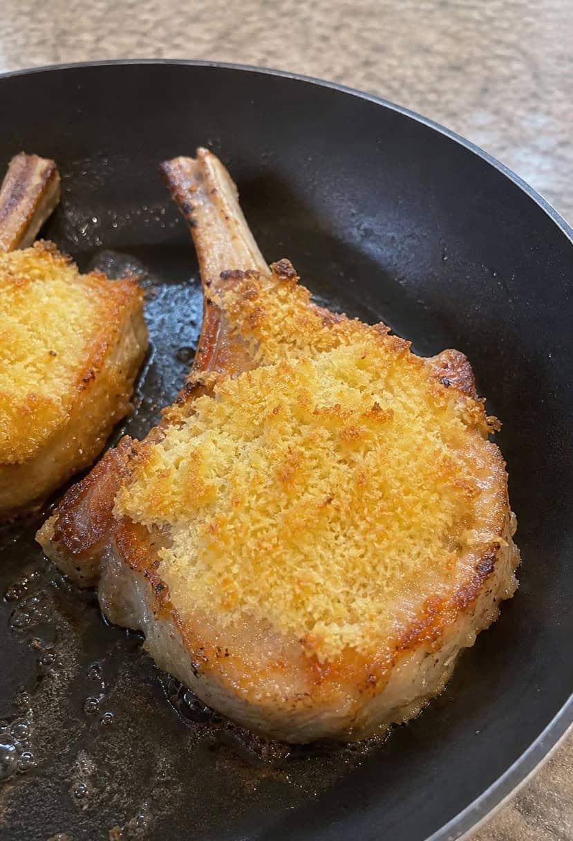 A golden-brown pork chop with a crispy parmesan crust sizzling in a skillet