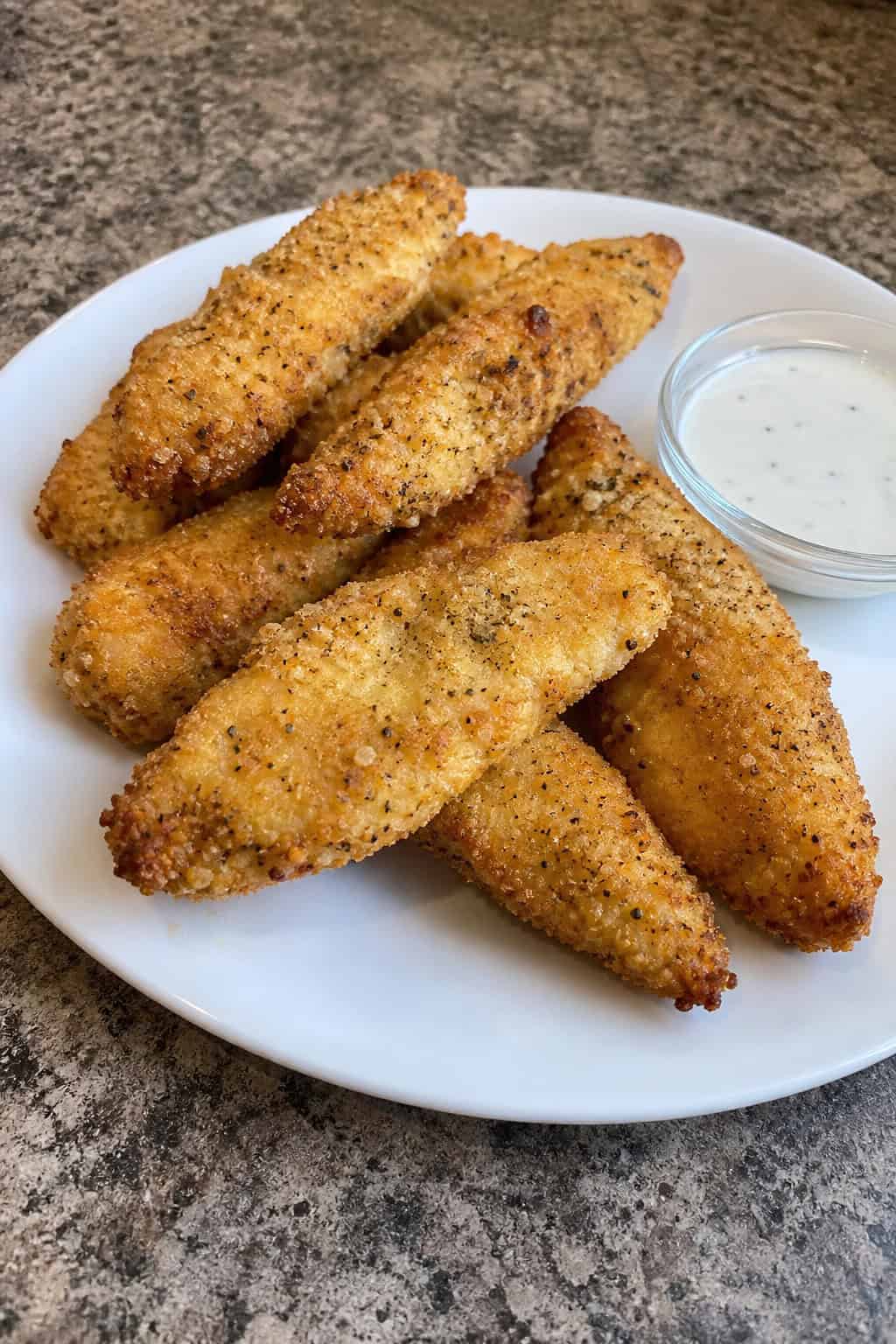 Crispy Parmesan garlic chicken strips on a wooden serving board, garnished with chopped parsley beside a small bowl of dipping sauce.