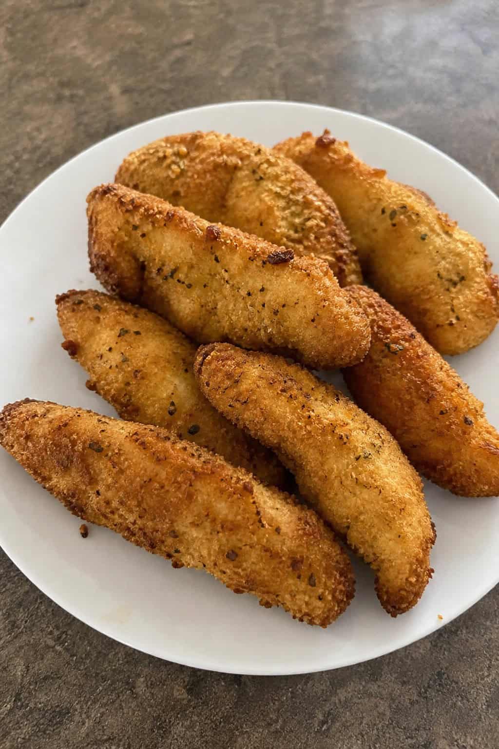 A plate of golden-brown Parmesan-herb chicken tenders garnished with fresh herbs and a bowl of dipping sauce on a kitchen table.