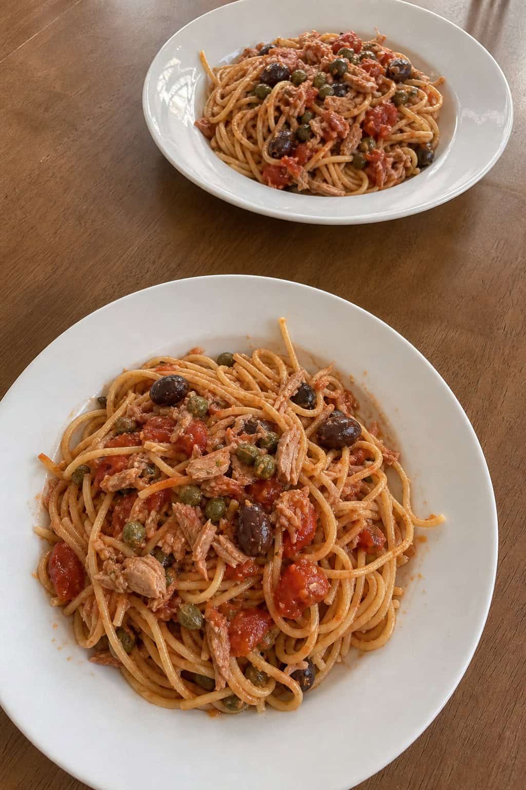 Two plates of pasta puttanesca with tomatoes, olives, and tuna on a wooden table set for two people.